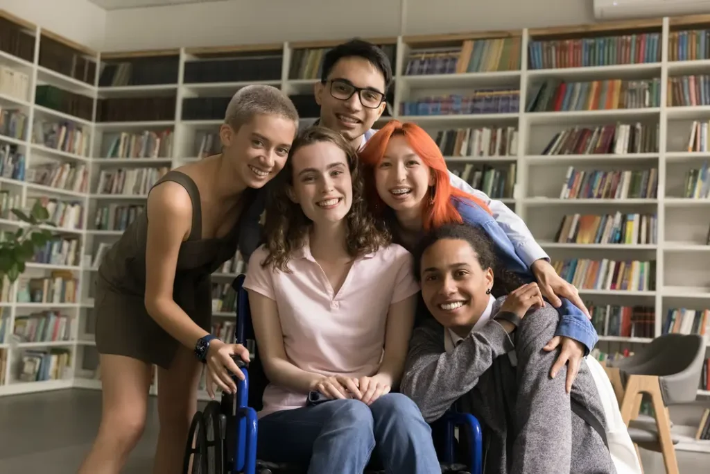 Group of young adults in a library huddled around a female participant in a wheelchair. They are all confidently smiling at the camera.